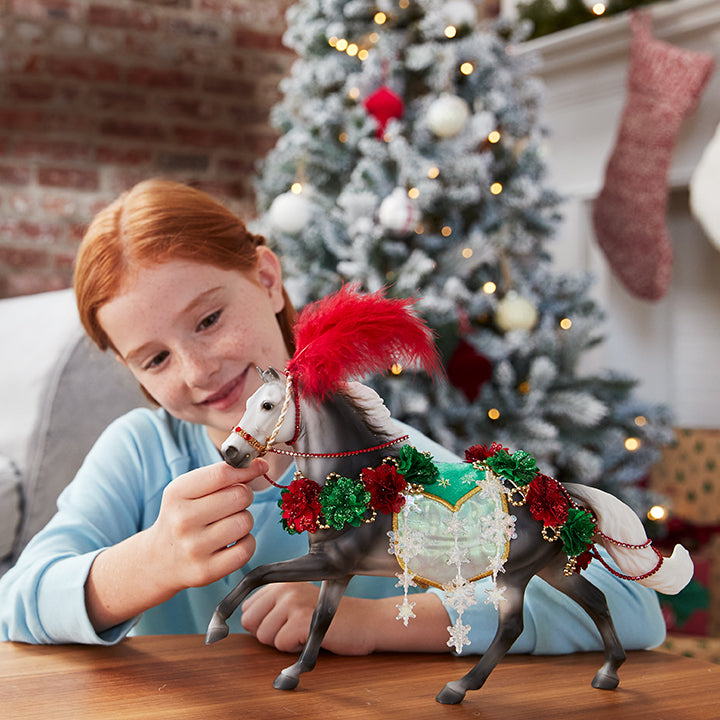 A child posing with Breyer's Holiday Horse on a table with a Christmas Tree in the background.