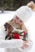 Girl wearing white beanie hat and sweater, holding the holiday horse, a bay warmblood, dress in a white blanket and wooden skis with a red bow